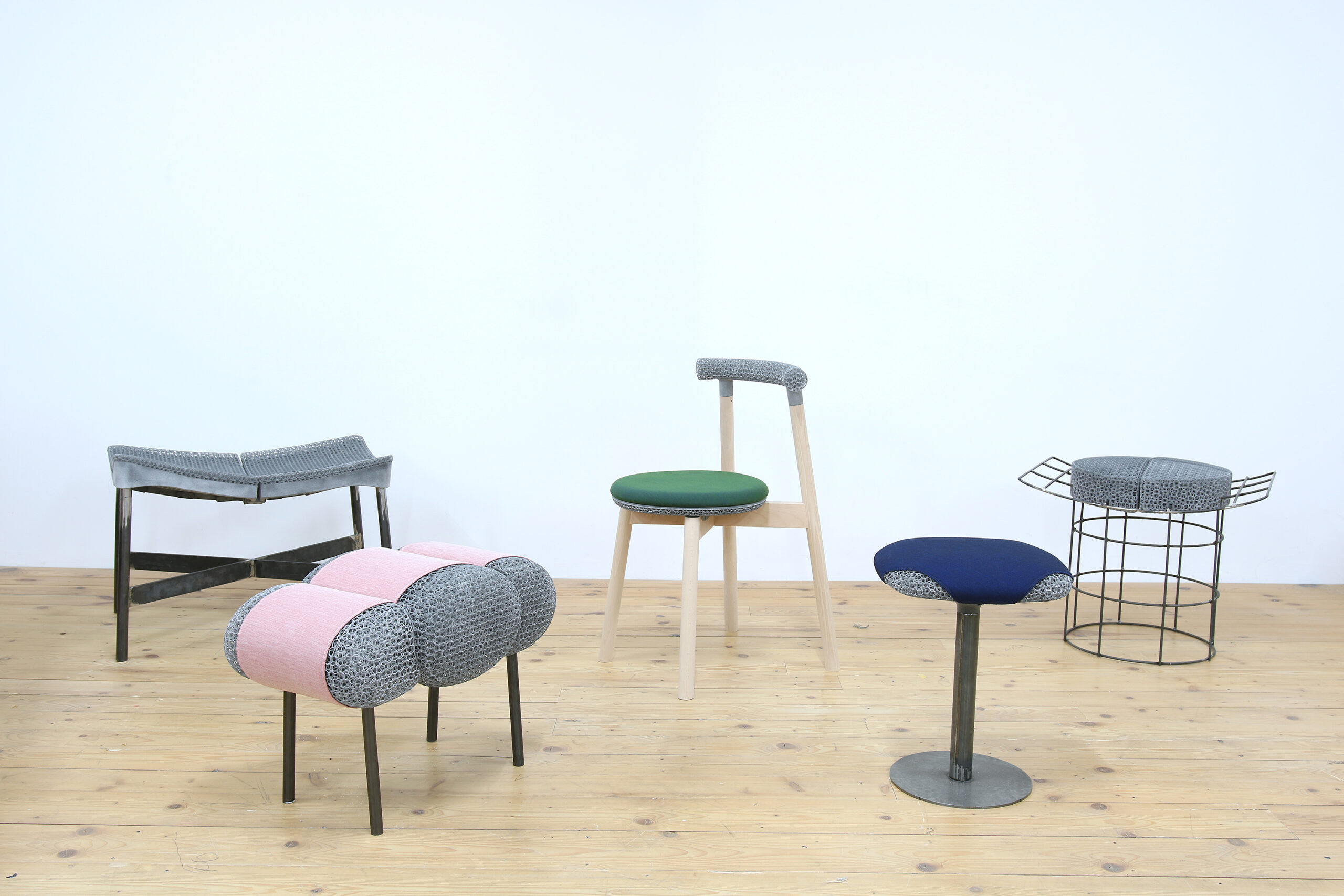 New possibilities of furniture production: Various chairs and stools in front of a white wall on a wooden floor with various substructures and 3D-printed seats and textile covers in blue, pink and green.