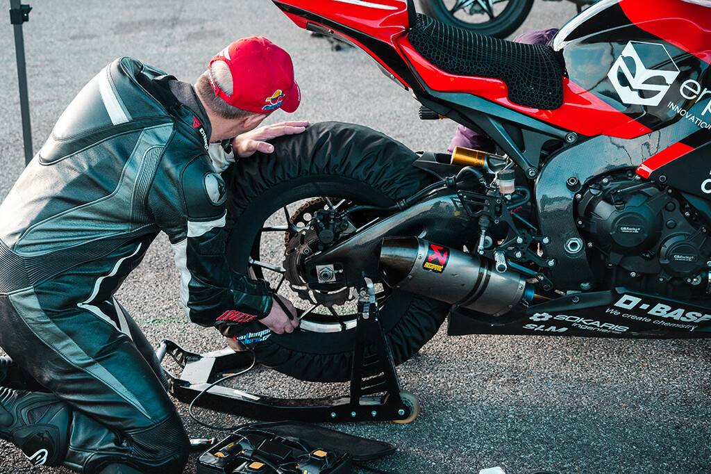 Slideshow of three photos: 1. A white man in motorcycle gear kneels in front of the rear wheel of his motorcycle that he has just changed. 2. A motorcyclist on a red and black motorcycle cornering at high speed. 3. Rear/top view of a red and black motorcycle and its custom seat with BASF logo.