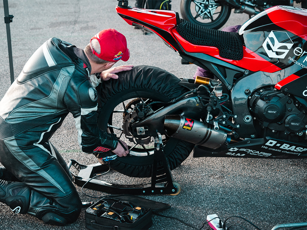 Slideshow of three photos: 1. A white man in motorcycle gear kneels in front of the rear wheel of his motorcycle that he has just changed. 2. A motorcyclist on a red and black motorcycle cornering at high speed. 3. Rear/top view of a red and black motorcycle and its custom seat with BASF logo.