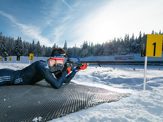 A young white biathlete surrounded by snow lying on the ground aiming his rifle at a target. Snow-covered fir trees in the background.