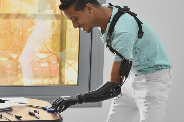 A young man of Color with short hair and a turquoise t-shirt wears a black prosthetic arm and reaches for a pair of scissors lying on a table.